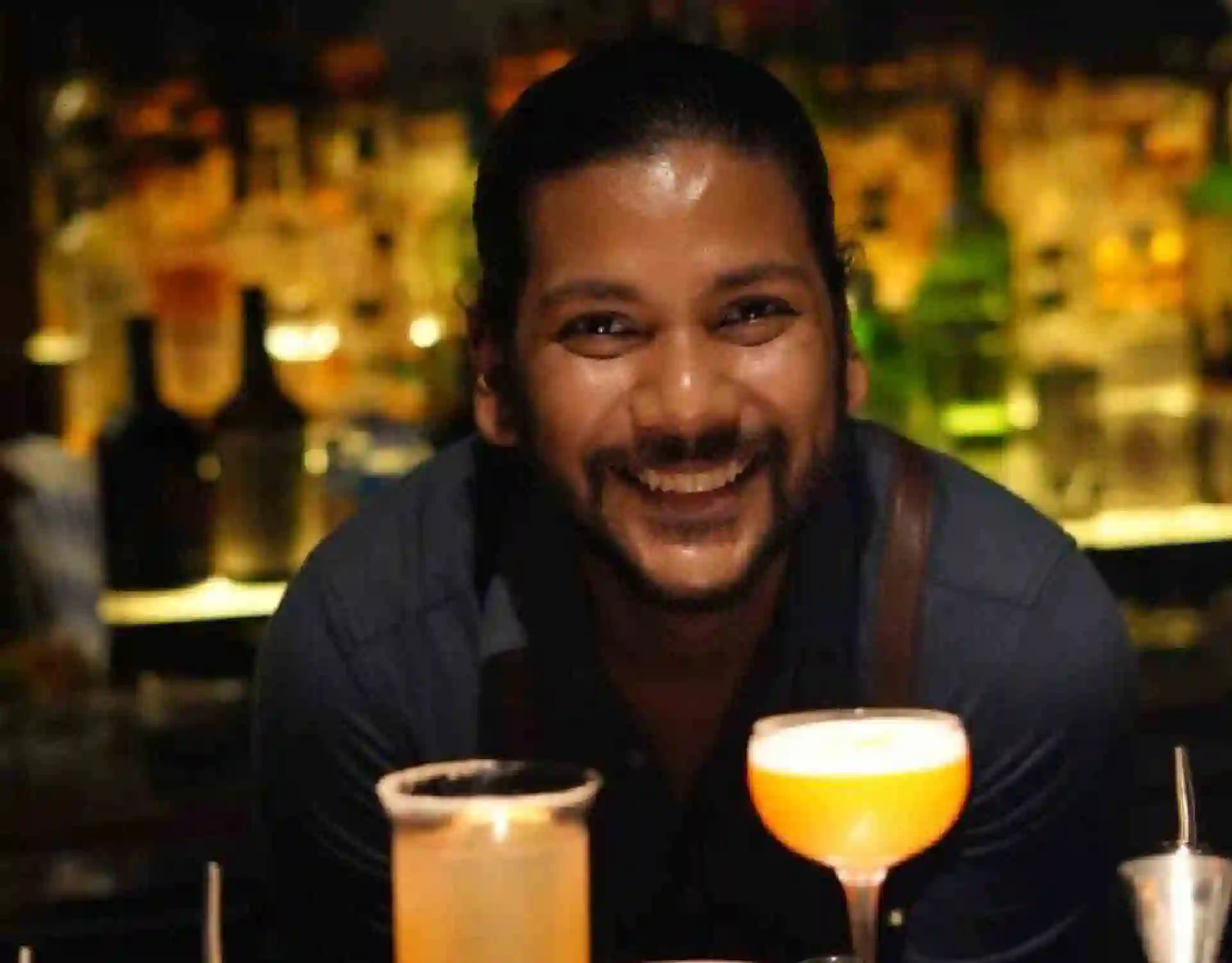 The bartender is seen smiling at the camera with cocktails displayed in front.