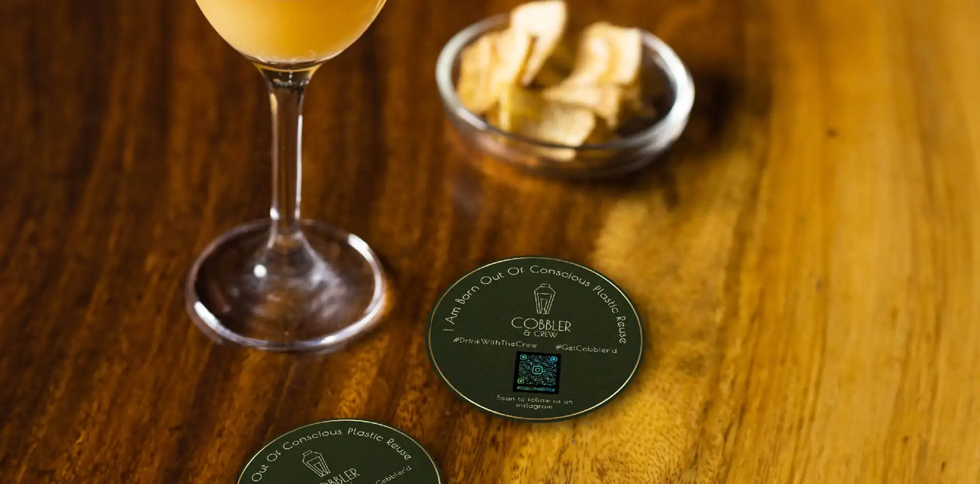 Cobbler & Crew coaster on a wooden table along with cocktail and chips.